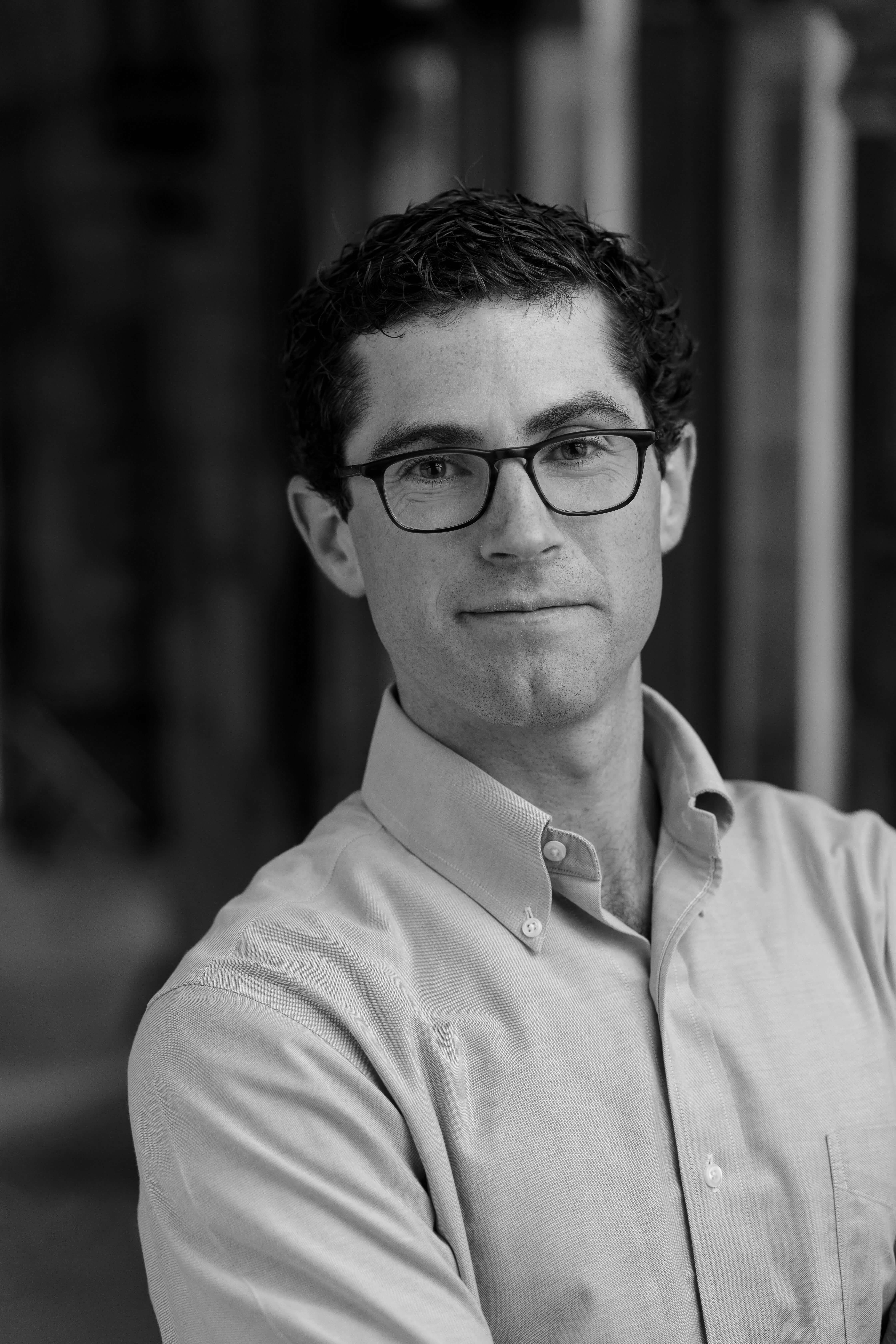 A black and white portrait photograph of a man with dark hair and glasses. He is wearing a light colored shirt and looking into the camera with a subtle smile.