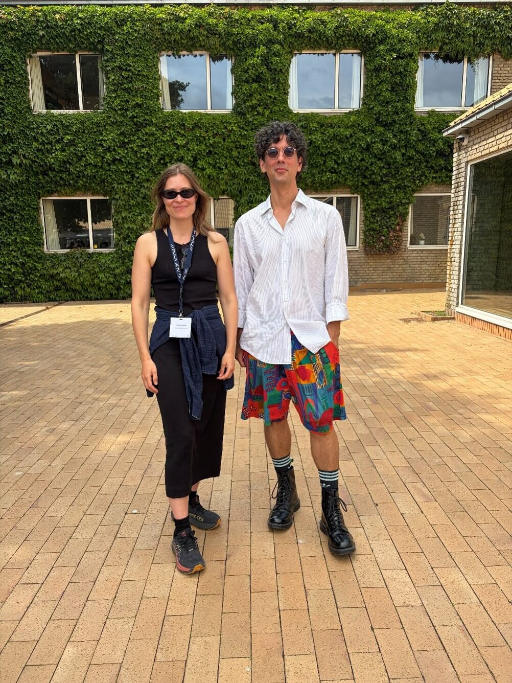 Two people standing next to each other outside. To the left a woman wearing a black skirt and tank top. To the right a man wearing colorful shorts and a button-up t-shirt. The woman has long straight hair and glasses. The man has shorter, dark, curly hair and glasses. They are smiling.