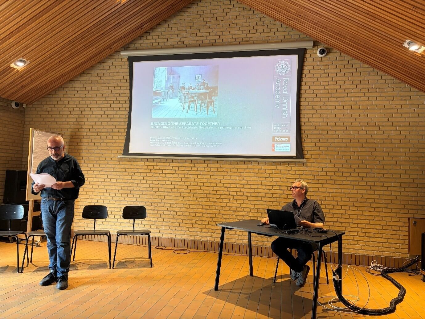 A man with glasses and short hair is standing in the left side of the photograph reading from a piece of paper. Another man with glasses is sitting at a desk in front of a laptop with a in the right side of the photograph. A big projector screen is behind the two men showing the title page of a presentation on Bindesbøll.